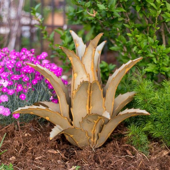 Metal agave sculpture placed in a garden with pink flowers and green bushes in the background.